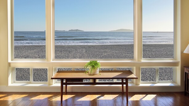A room with a large window overlooking a beach, a wooden table, and a potted plant.