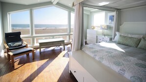 A bedroom with a large window overlooking a beach, a bed with a patterned bedspread, a chair with a cushion, and a nightstand with a lamp.
