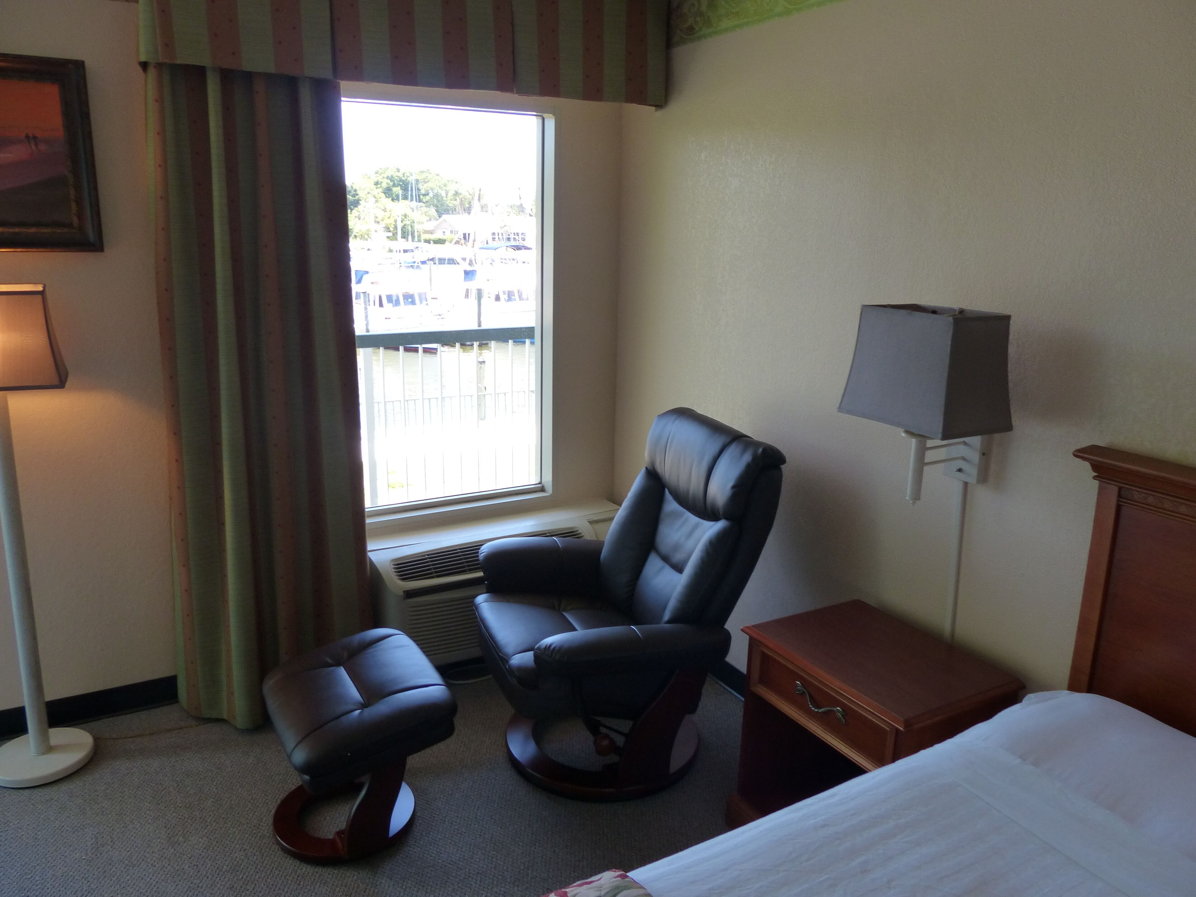 A hotel room with a bed, a desk, a chair, a lamp, a window with curtains, and a view of the outside.