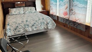A bedroom with a wooden paneled wall, a bed with floral bedding, a nightstand, a chair, and a window with a seascape mural. - Whale Cove suites (Whale Cove)