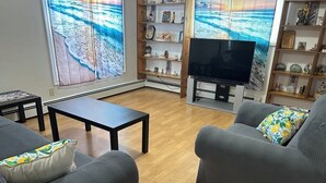A living room with two grey sofas, a coffee table, a flat-screen TV, and a bookshelf with various items. - Whale Cove suites (Whale Cove)
