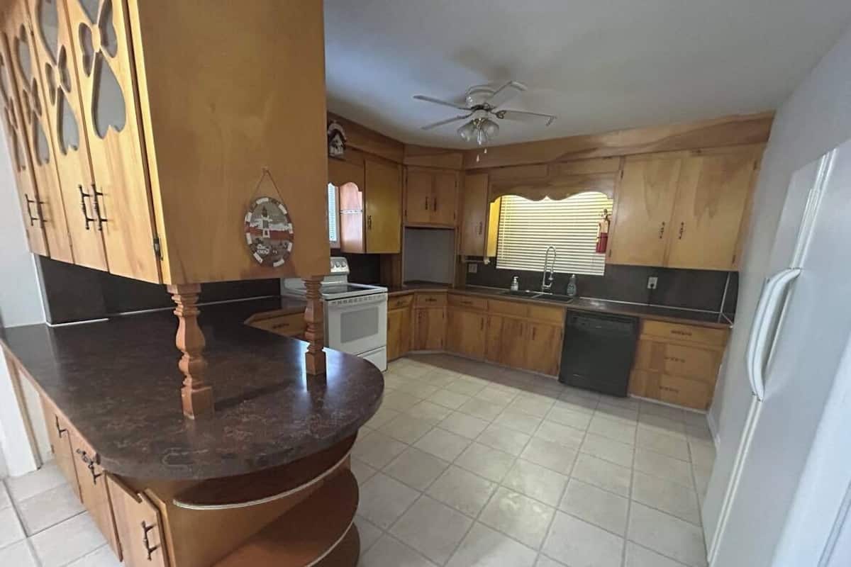 Una cocina con armarios de madera, encimera oscura, ventilador de techo y ventana con persianas.