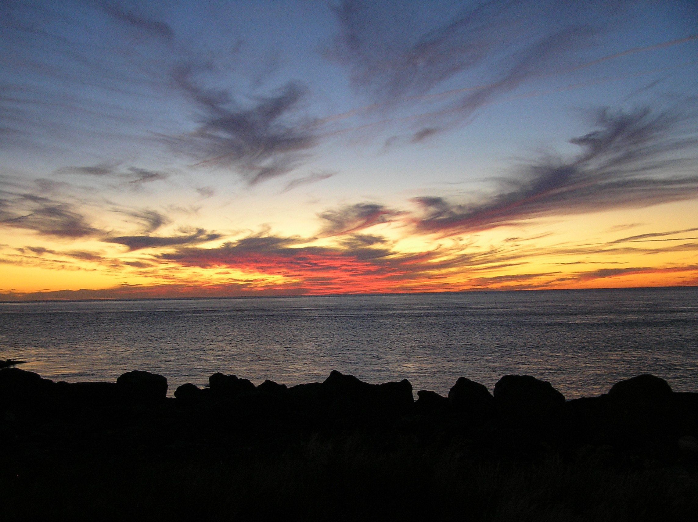 A sunset over a calm sea with rocky coastline silhouetted in the foreground.