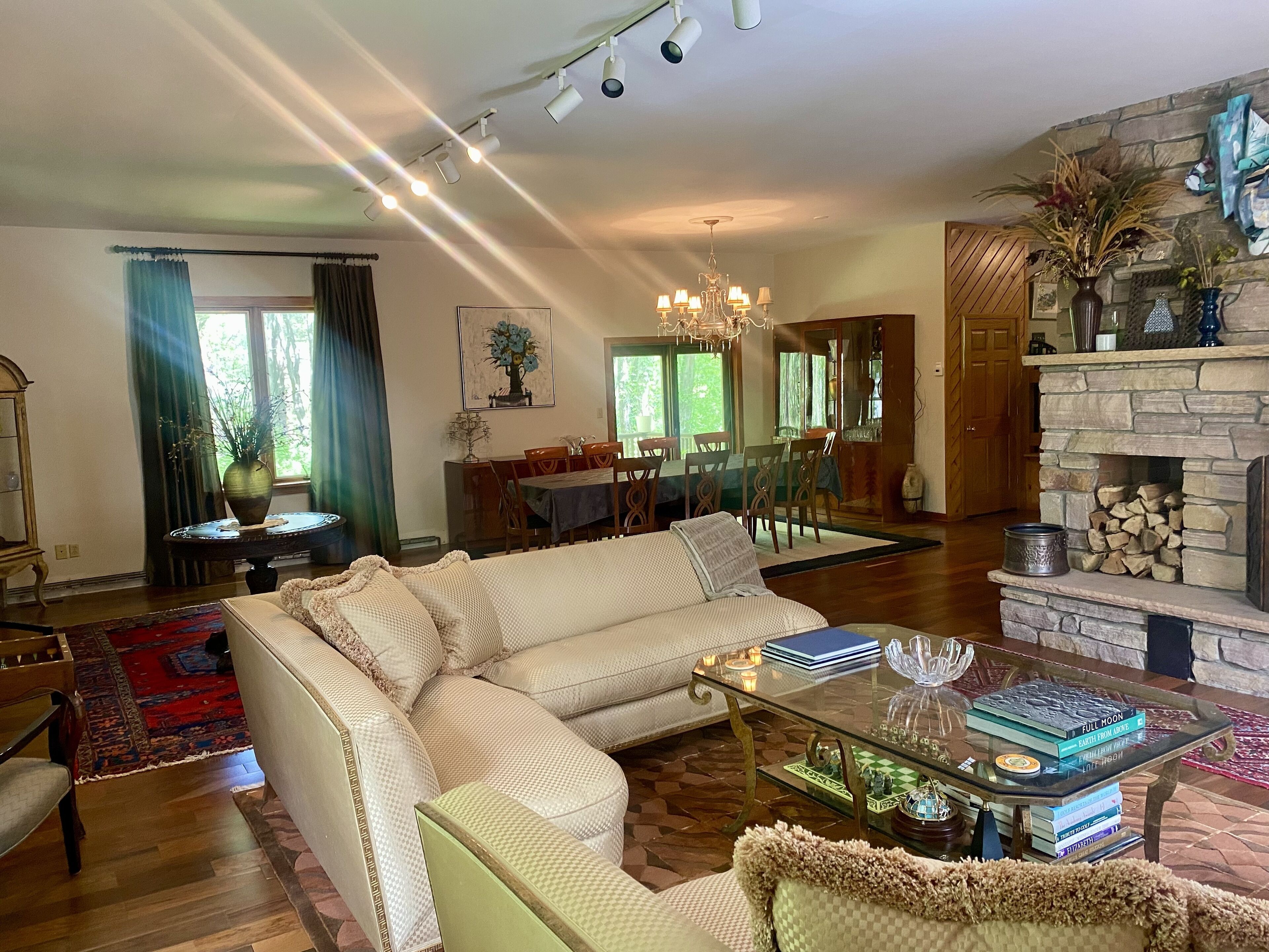 A spacious living room with a large sectional sofa, a glass coffee table, and a fireplace. There is a dining area in the background with a chandelier.