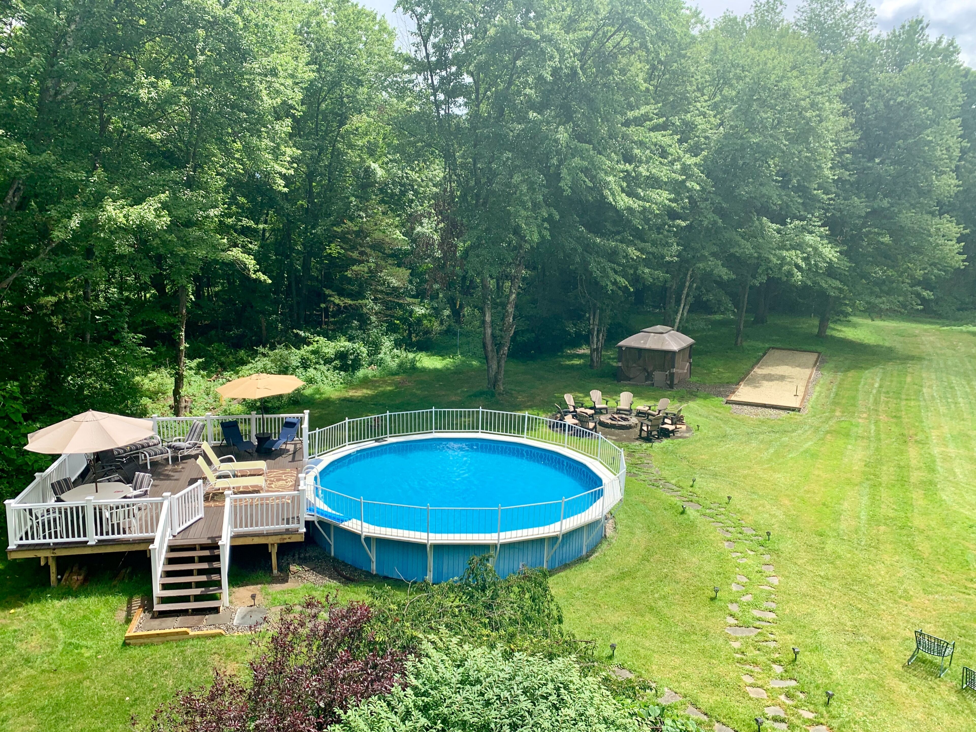 A circular pool with a wooden deck and a white railing, surrounded by a well-maintained lawn and trees.