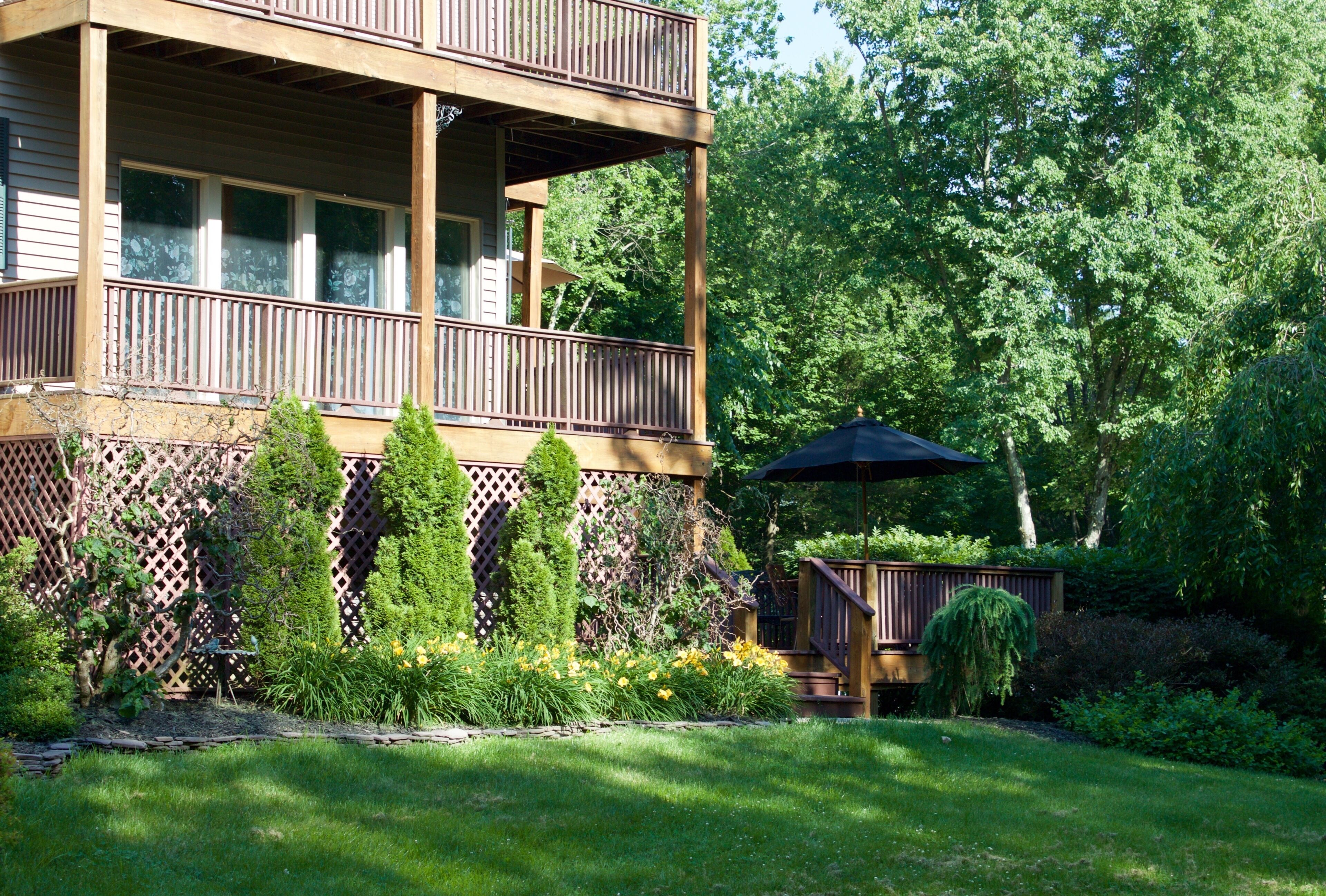 Une maison en bois à deux étages, dotée d’un balcon, entourée d’une végétation luxuriante et d’un jardin bien entretenu.