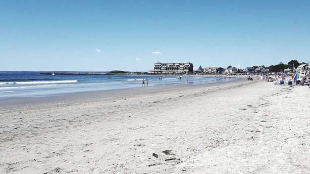Ein Strand mit einer weiten Sandfläche, wenigen Spaziergängern und Gebäuden im Hintergrund.