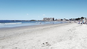 A beach with a long stretch of sand, a few people walking, and buildings in the background.