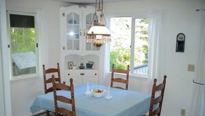 A dining room with a table set for four, wooden chairs, a chandelier, and a view of greenery outside. - Bass Harbor Oceanfront Chalet:  Peaceful, Private, Great On-The-Ocean View (Bass Harbor)