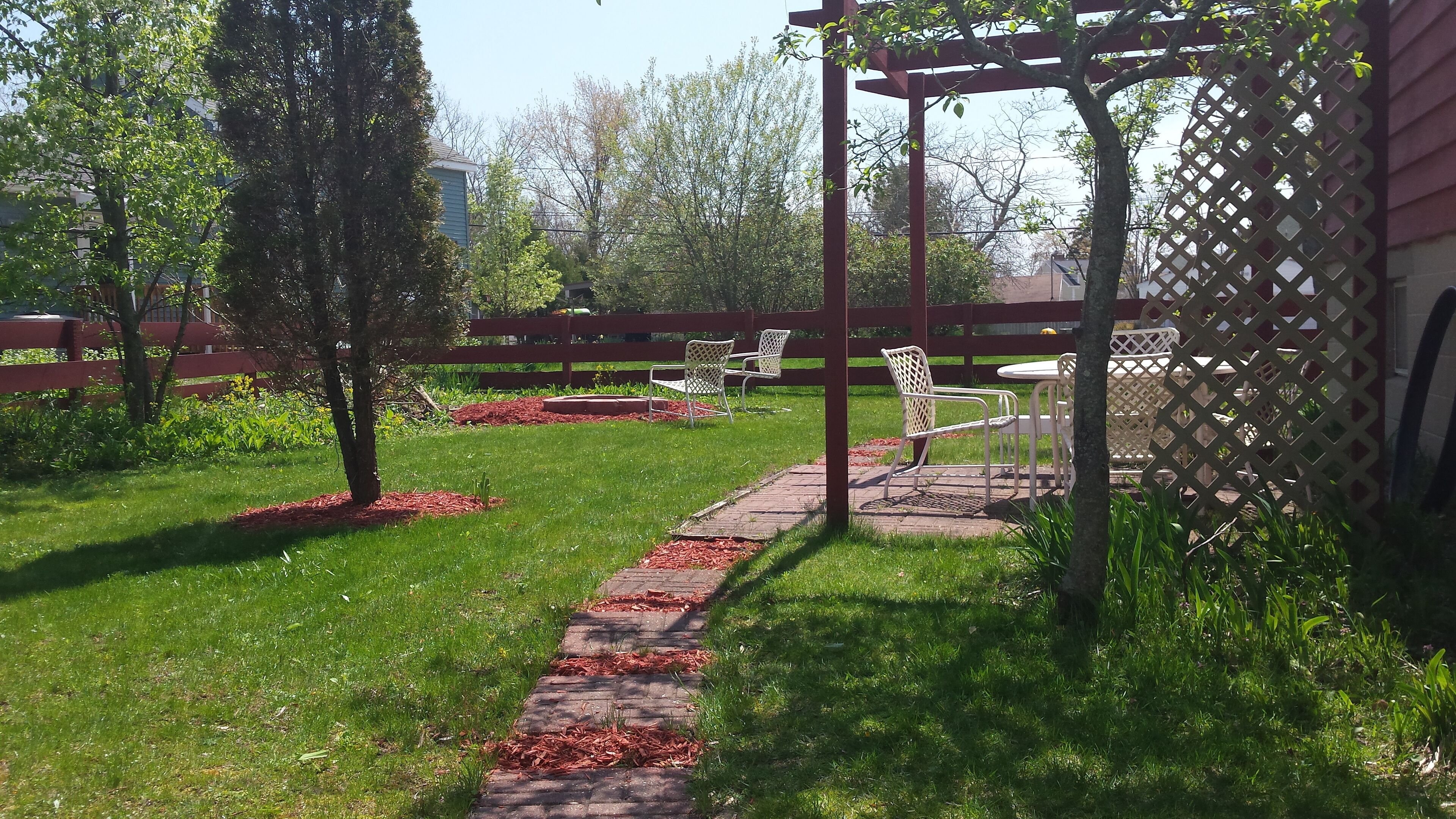 Ein Garten mit gepflastertem Weg, Metallstühlen und einer Pergola.