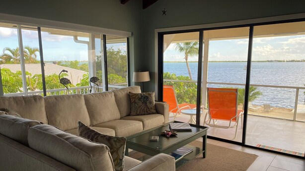 Ein Wohnzimmer mit einem beige farbenen Sofa, einem Glastisch und Blick auf Strand und Meer durch große Fenster.