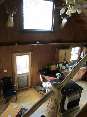 Interior rústico de uma cabana, com área de jantar, cozinha e uma grande janela que proporciona luz natural.