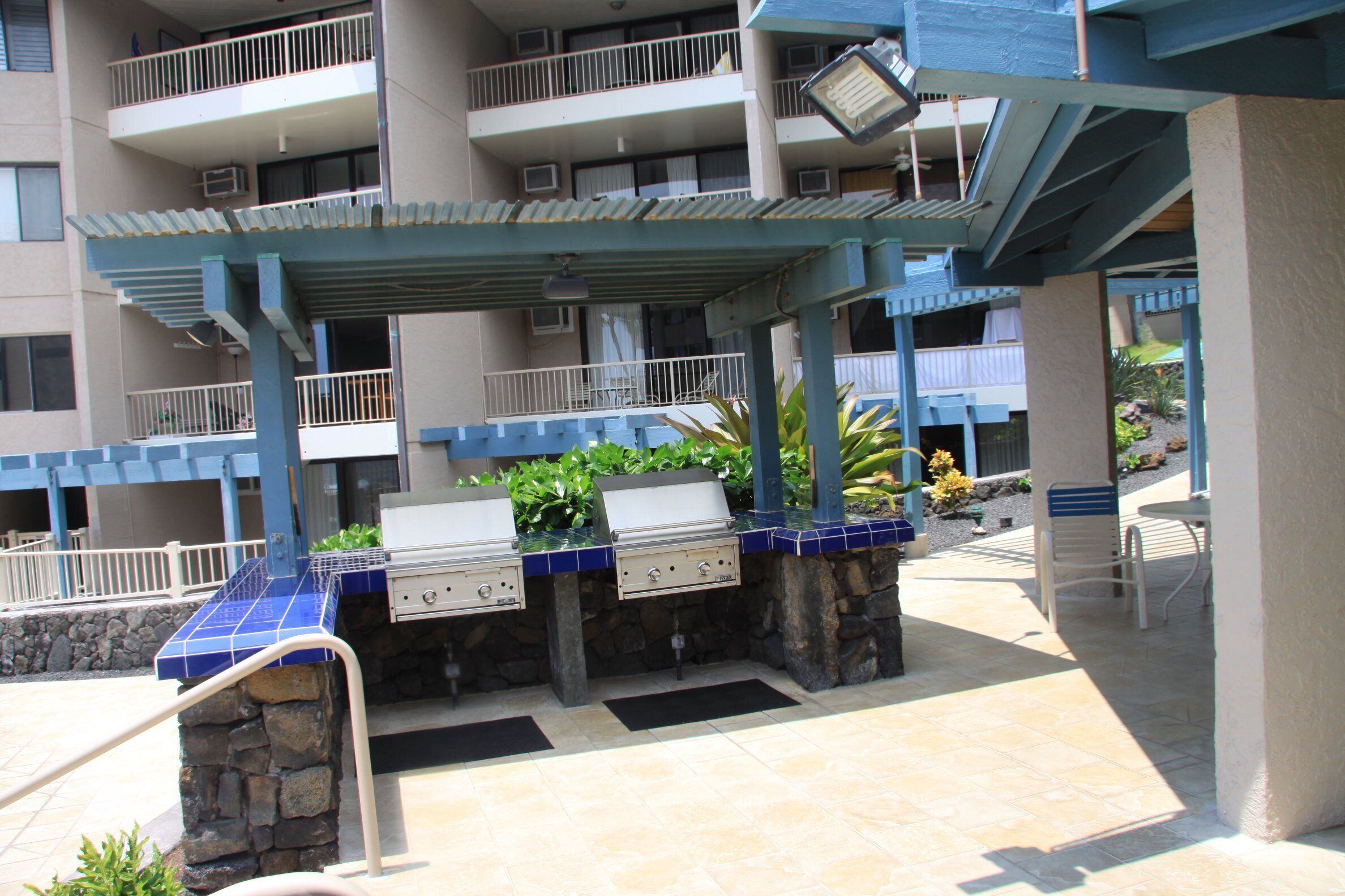 An outdoor barbecue area with a stone wall and grill, surrounded by a covered patio with blue tiles and a white railing.