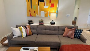 Un salon moderne avec un canapé d’angle gris, une table basse en bois et un plaid rouge. Un tableau décoratif est accroché au mur au-dessus du canapé, et une lampe se trouve sur une table d’appoint.