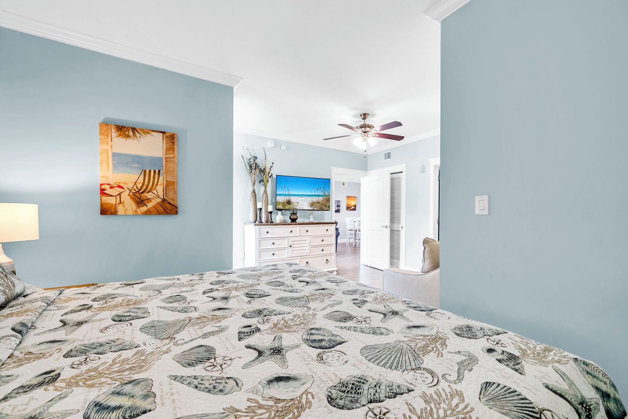 A bedroom with a beach-themed bedspread, a ceiling fan, and a painting of a beach scene.