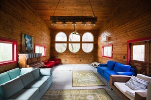 Interno di una baita di legno con un divano blu e bianco, una poltrona rossa, un pianoforte in legno e un'ampia finestra con vista sugli alberi.