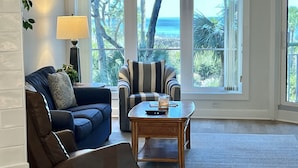 Un vaste salon doté d’une grande fenêtre offrant une vue sur l’océan et les palmiers. On y trouve un canapé bleu, un fauteuil à rayures et une table basse en bois.