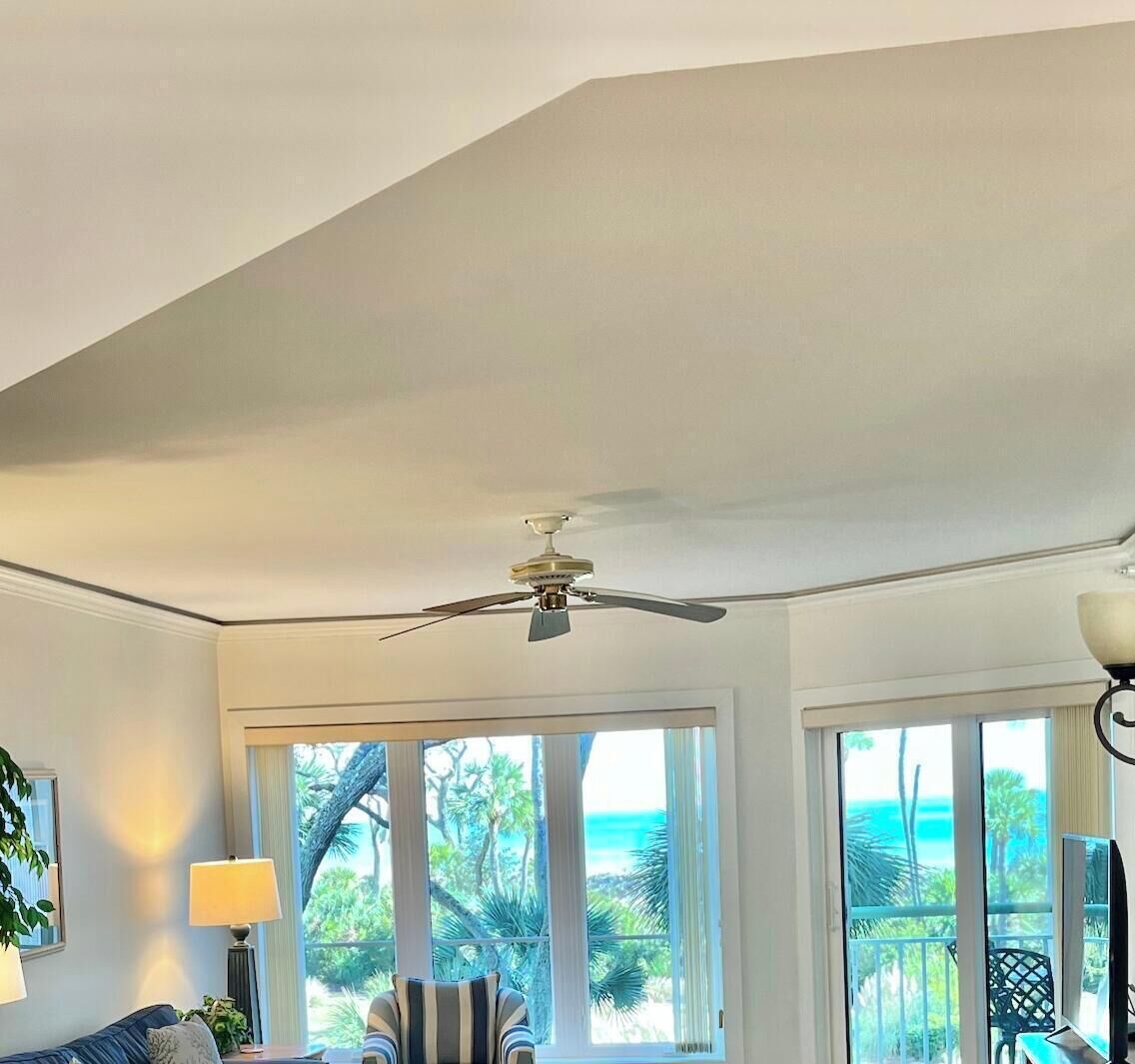A room with a ceiling fan, large windows with a view of the ocean, a sofa, a chair, and a lamp.