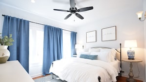 Quarto com cama, ventilador de teto, mesa de cabeceira com abajur e janela com cortinas.
