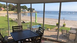 A covered patio with a table and chairs, overlooking a beach with boats and a pier.