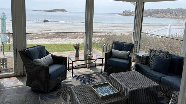 A sunlit room with wicker furniture, large windows offering a view of the beach and ocean.