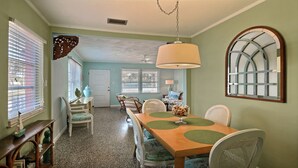 A dining area with a long wooden table, green placemats, and white chairs. There is a large window with blinds, a door, and a hanging light fixture. - SOUTH BEACH & PARK Just a Hop Skip & Jump • CORAL COTTAGE • Fun Seaside Vero  (Vero Beach)