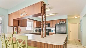 A kitchen with wooden cabinets, a central island, and a dining area with green chairs. - SOUTH BEACH & PARK Just a Hop Skip & Jump • CORAL COTTAGE • Fun Seaside Vero  (Vero Beach)