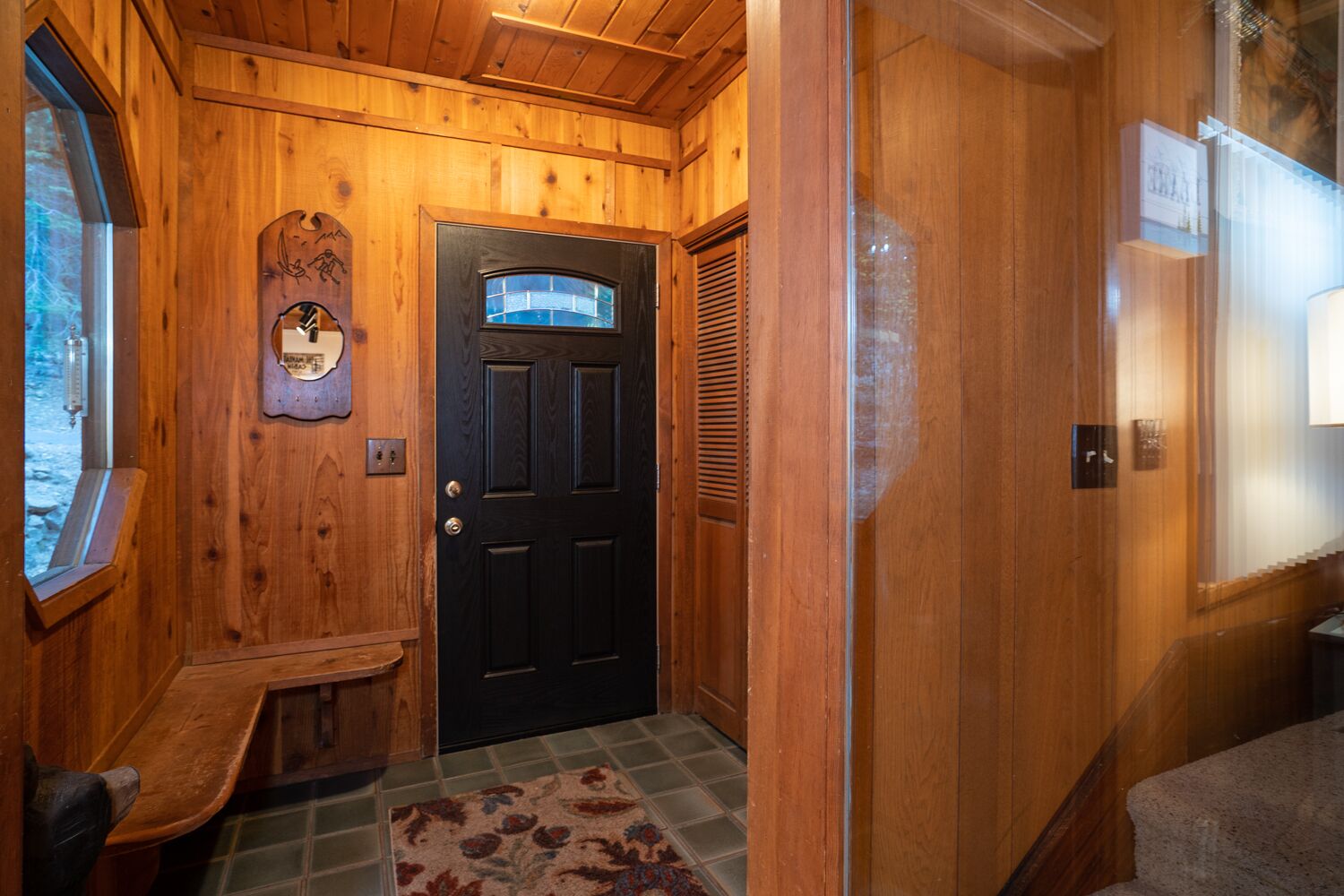 A wooden interior hallway with a black door, a bench, a wall-mounted thermometer, and a patterned rug.