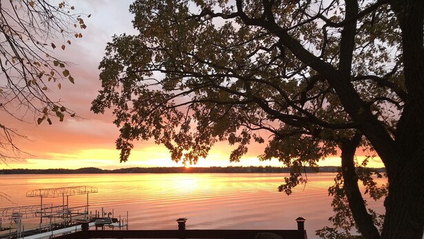 A sunset view from a deck with wicker furniture and a railing overlooking a calm body of water.