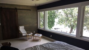 A bedroom with a bed, a chair, a small table with a laptop, and a large window with a view of trees and a parking lot.