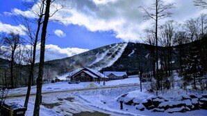 A mountain lodge with a snow-covered landscape, a clear sky, and a visible chimney emitting smoke.