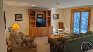 A living room with a wooden entertainment center, a television, a lamp, a sofa, a chair, and a table with a chess set.