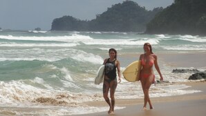 Zwei Personen laufen mit Surfbrettern am Strand entlang, im Hintergrund brechende Wellen und in der Ferne ein bewaldeter Bereich.