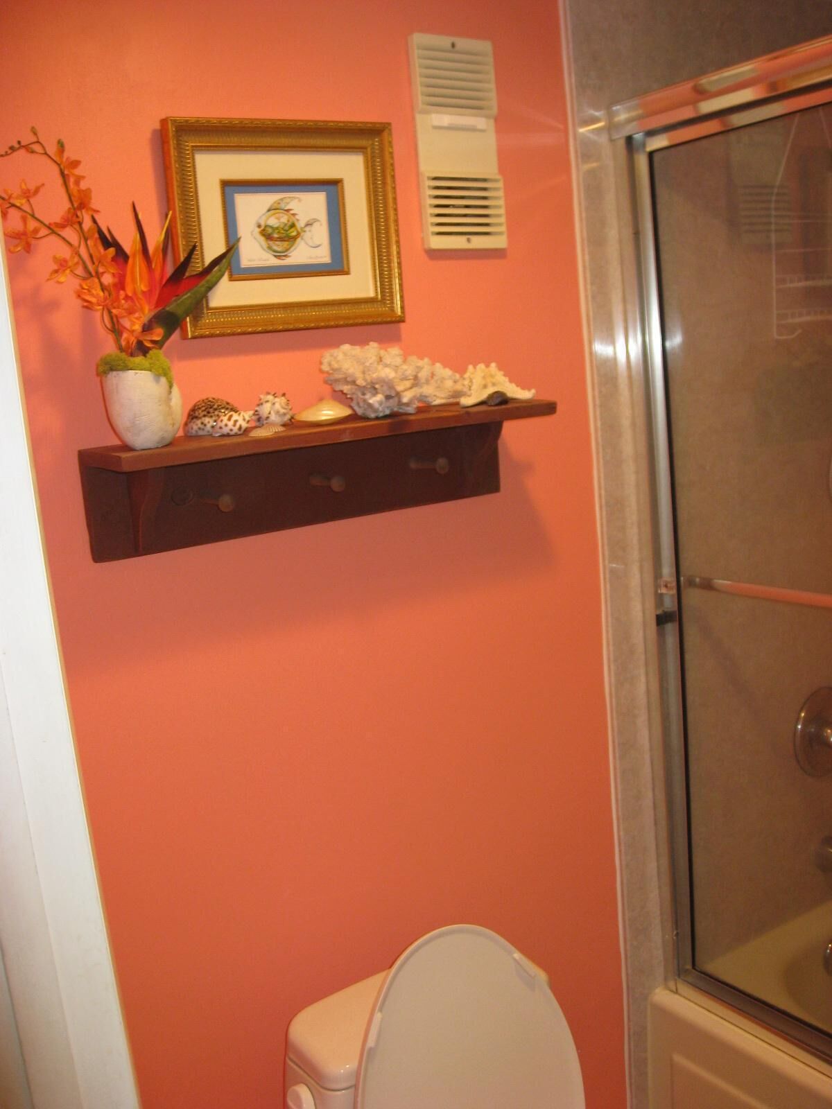 A bathroom with a toilet, a shower enclosure, a shelf with decorative items, and a framed picture on the wall.