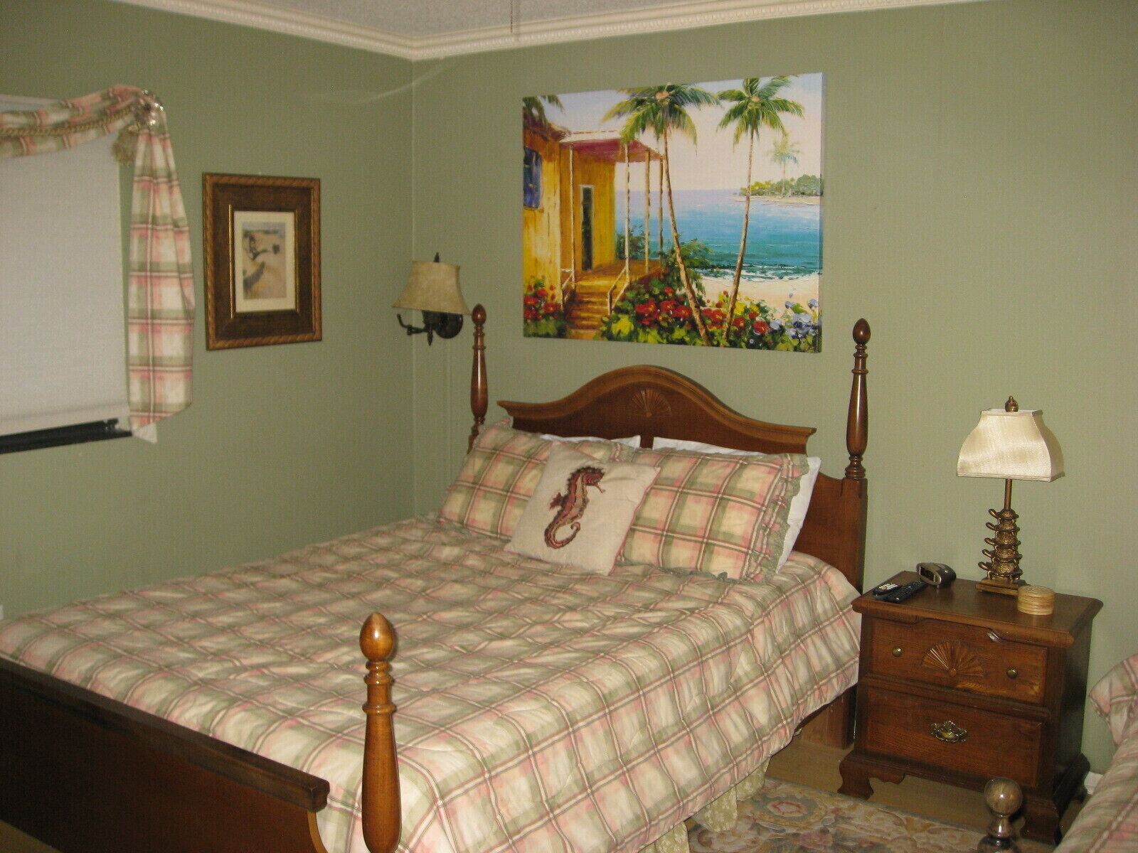 A bedroom with a bed, a nightstand, a lamp, a painting of a tropical scene, and a plaid curtain.
