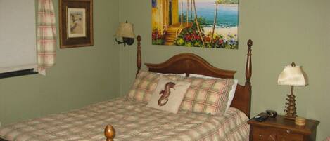 A bedroom with a bed, a nightstand, a lamp, a painting of a tropical scene, and a plaid curtain.