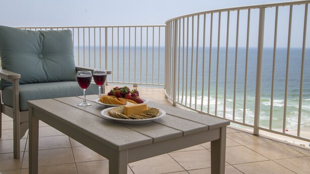 A balcony with a table set for two, featuring a plate of fruit and cheese, two glasses of red wine, and a view of the ocean. - 4 bedroom D Tower!! Turquoise Complex (Orange Beach)