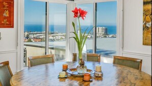 A dining area with a round table, six chairs, a vase with a red flower, and a view of the ocean through large windows. - Stunning Oceanview Craftsman in the village Of La Jolla - Walk To All! (San Diego)