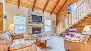 A cozy living room with leather sofas, a stone fireplace, a flat-screen TV, and a wooden staircase.