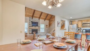 A spacious dining area with a long wooden table set for four, a stone fireplace, and a kitchen with wooden cabinets and modern appliances.
