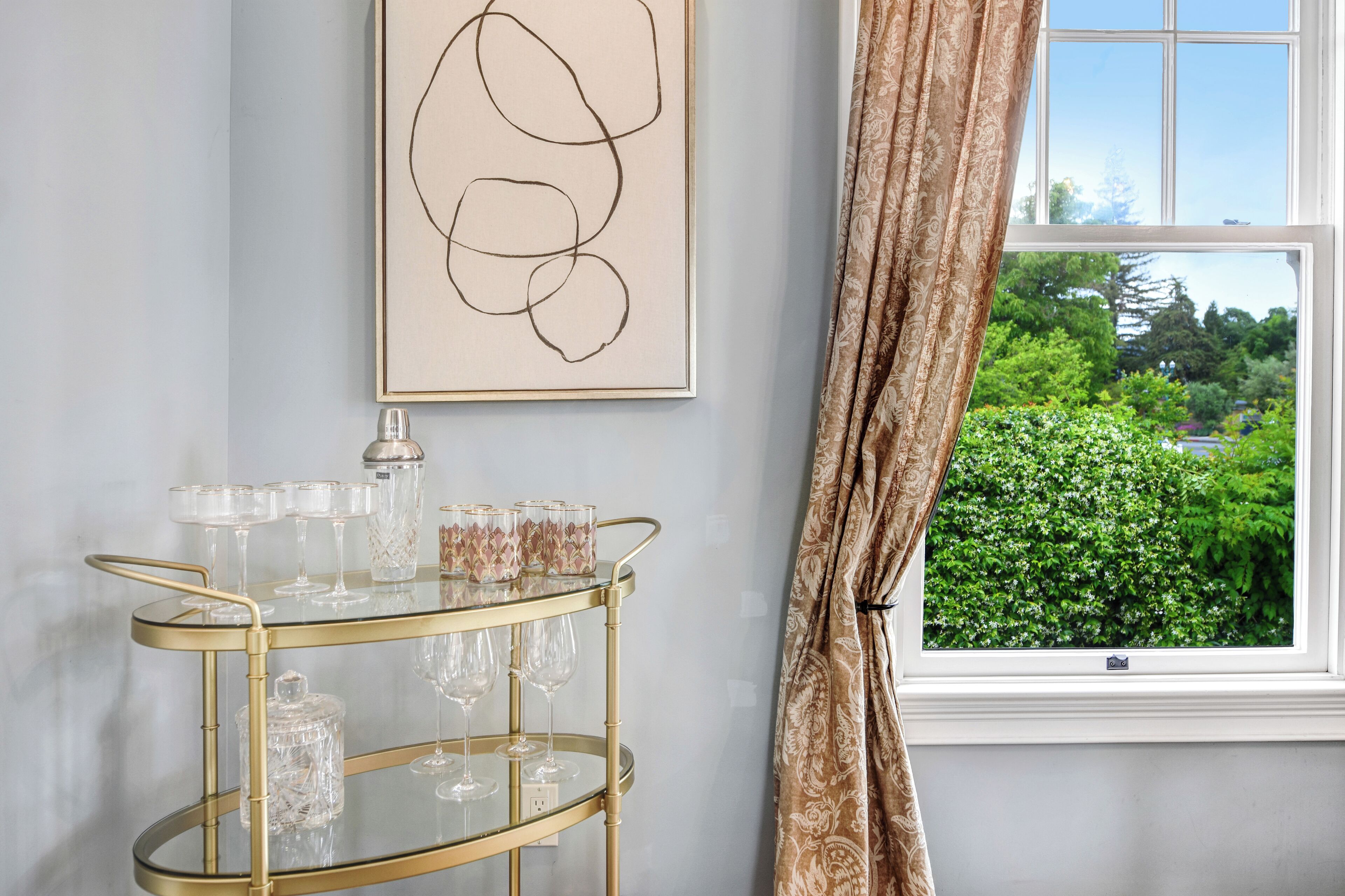 A room with a gold-framed glass cart holding wine glasses and a crystal decanter, a framed abstract artwork on the wall, and a window with a view of greenery.