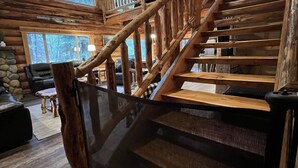 Eine Holztreppe mit Glastrennwand, ein Steinkamin und Blick auf den Wald durch große Fenster.