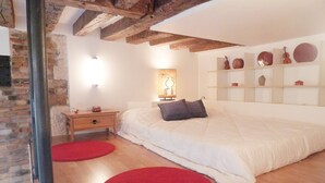 Un dormitorio con una cama grande, vigas de madera, pared de ladrillo, una mesita de noche, una alfombra roja y platos decorativos en estantes.