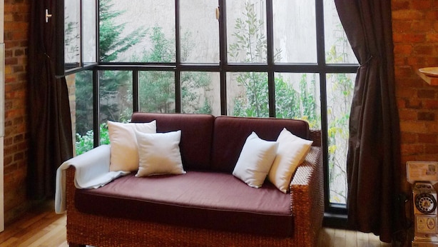 Una habitación con un ventanal grande, un sofá de mimbre con cojines, suelo de madera y pared de ladrillo.