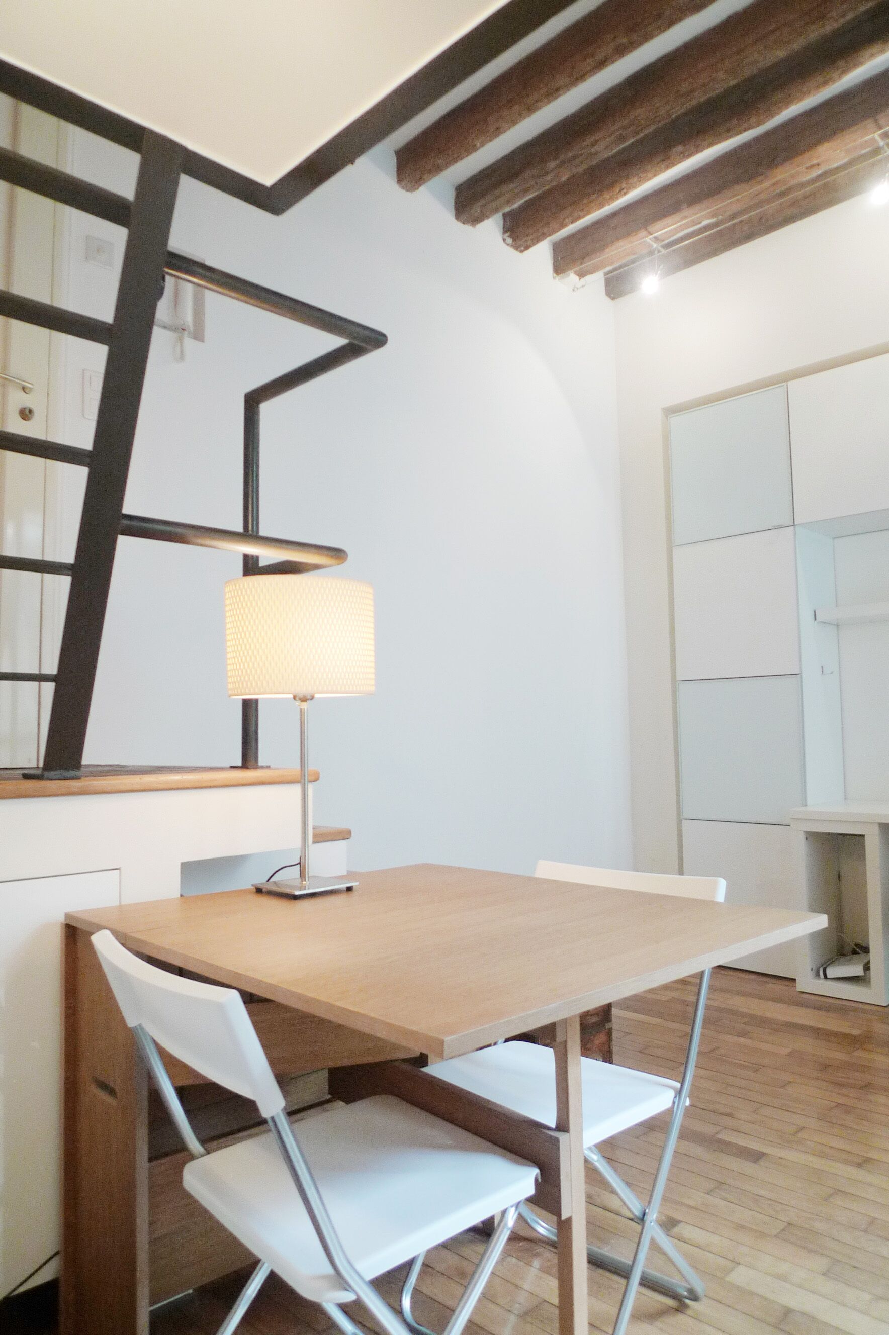 Un'angusta zona pranzo con un tavolo in legno e due sedie bianche, una lampada e una scala con travi a vista.