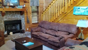 Ein gemütliches Blockhaushaus-Interieur mit Kamin aus Stein, Holzwänden, einem braunen Sofa, einem Couchtisch und einer Treppe.