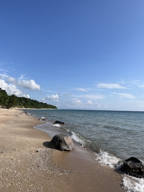 Una playa con piedras y rocas, cielo despejado y un bosque lejano.