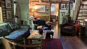 A cozy cabin interior with a wooden ceiling, a fireplace, a sofa, a chair, and a dining table.