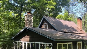 A rustic cabin with a stone chimney, large windows, and surrounded by lush greenery.