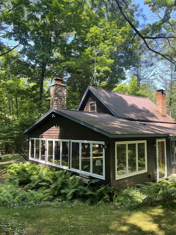 Una cabaña rústica con chimenea de piedra, ventanas amplias y rodeada de exuberante vegetación.